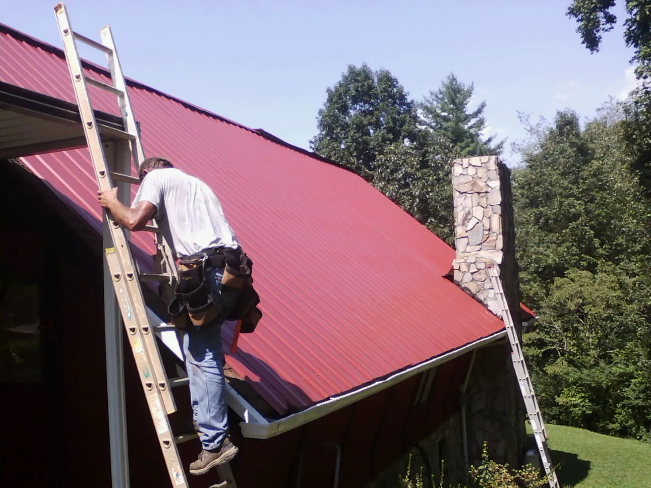 Roofer on a ladder beside a red metal roof and stone chimney — Soffit & Fascia Repair by Roofing Brothers in Mont Clare